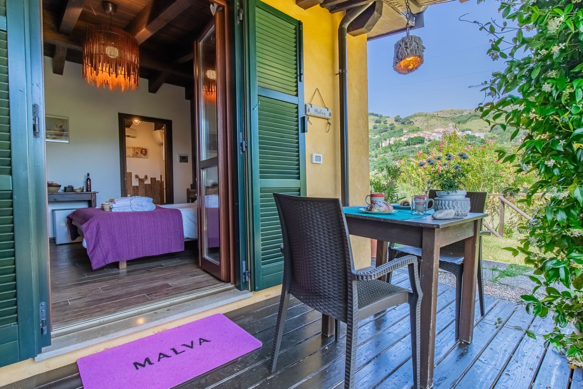 A room with a bed, a table with chairs, a hanging light fixture, and a view of greenery and hills.