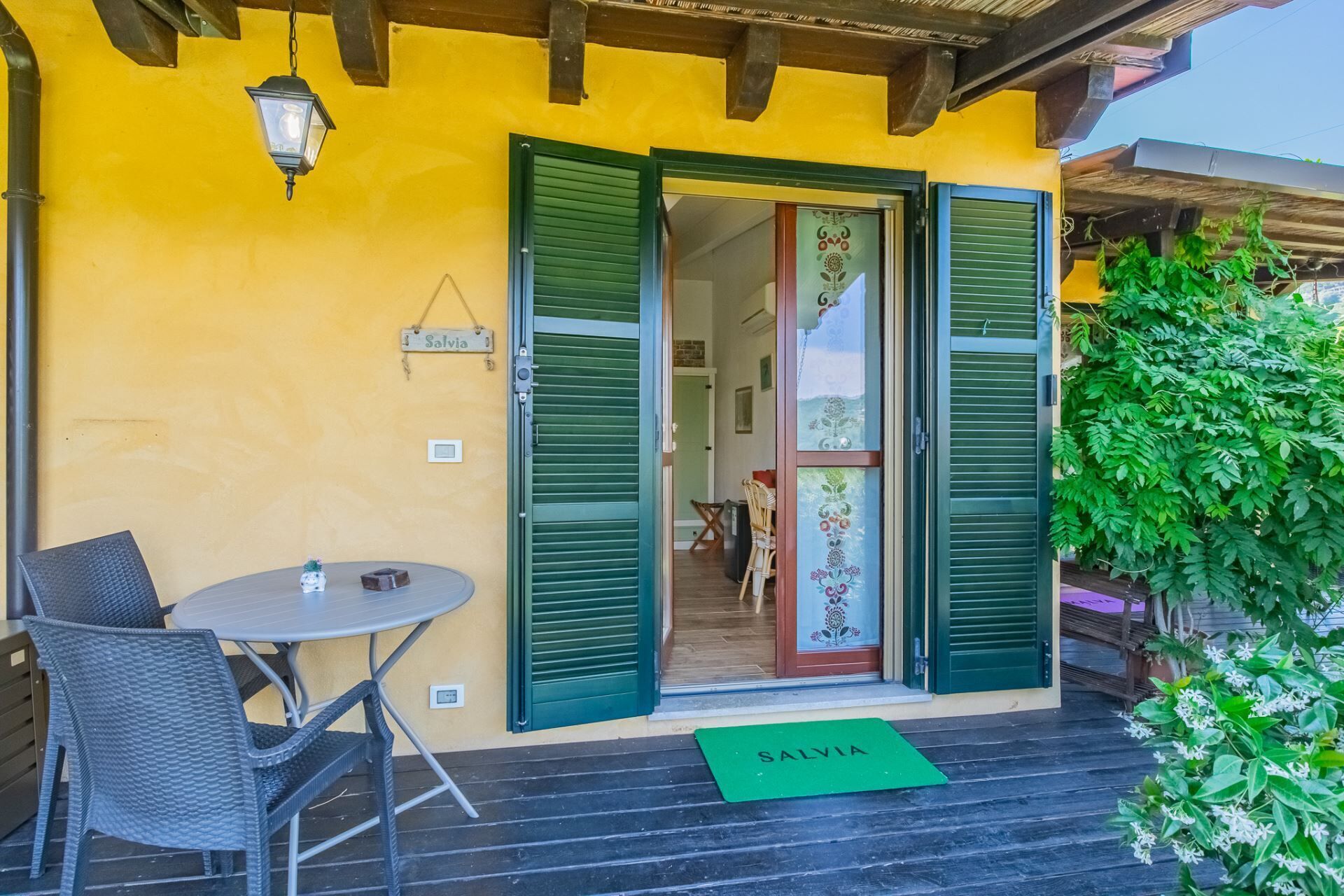 A yellow building with green shutters, a table with chairs, and a green mat with 'SALVIA' on it.