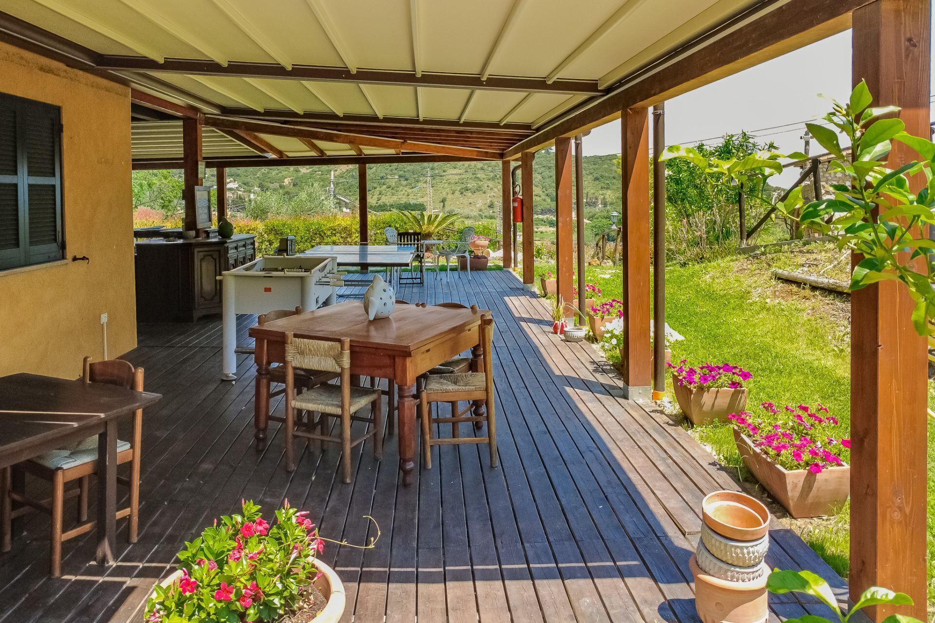 Een terras buiten met een houten dek, een tafel met stoelen en potplanten.