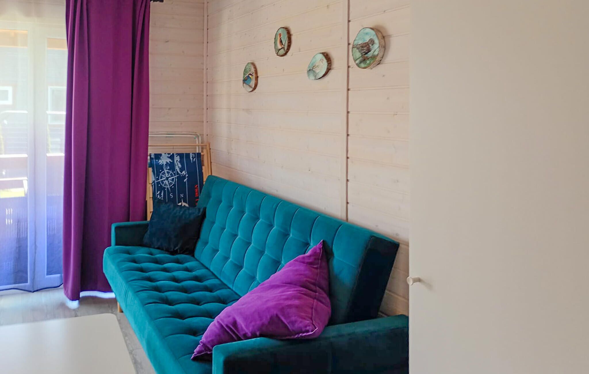 A teal sofa with a purple pillow, a compass wall hanging, and a window with purple curtains.