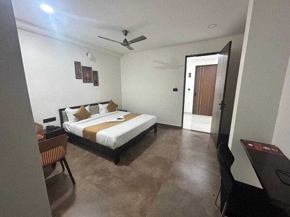 A hotel room with a bed, a desk, a chair, a ceiling fan, and a door leading to another room. - Classic Adithya Stay (Hyderabad)