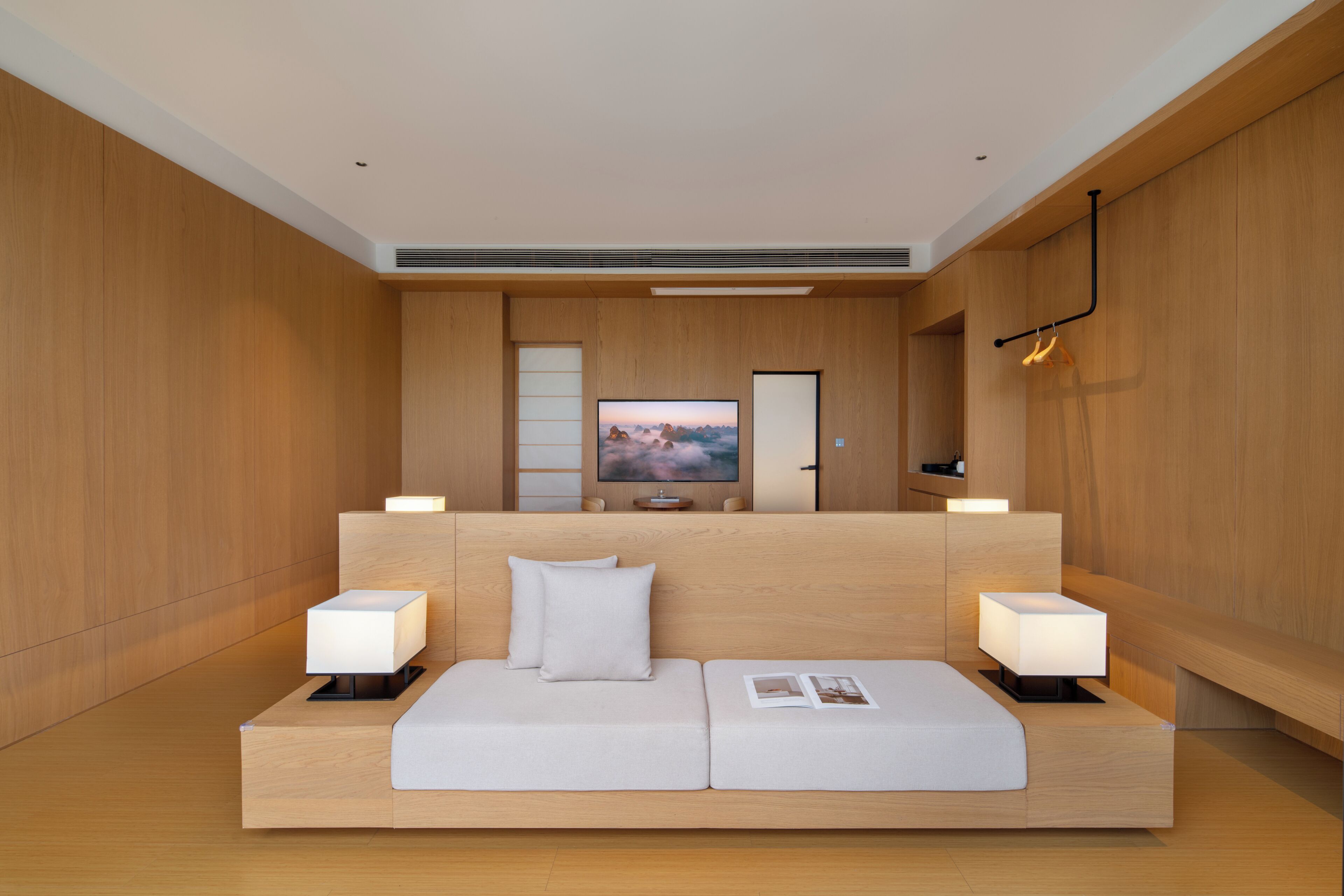 A modern hotel room with a wooden panel wall, a flat-screen TV mounted above a sofa, and two bedside tables with lamps.