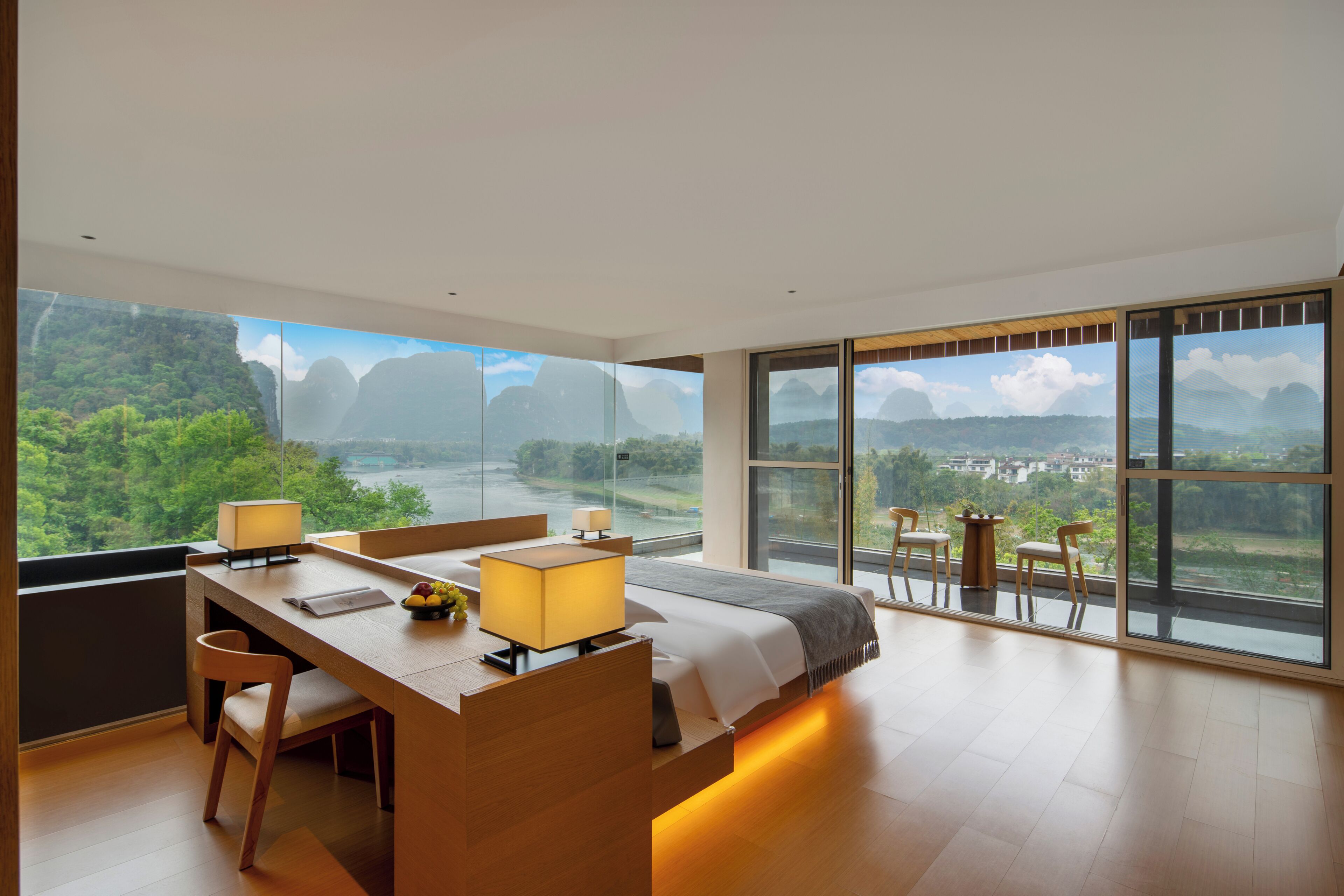 Ein modernes Hotelzimmer mit einem großen Bett, einem Schreibtisch mit Stuhl, einem kleinen Tisch mit Lampe und Blick auf eine gebirgige Landschaft durch große Fenster.