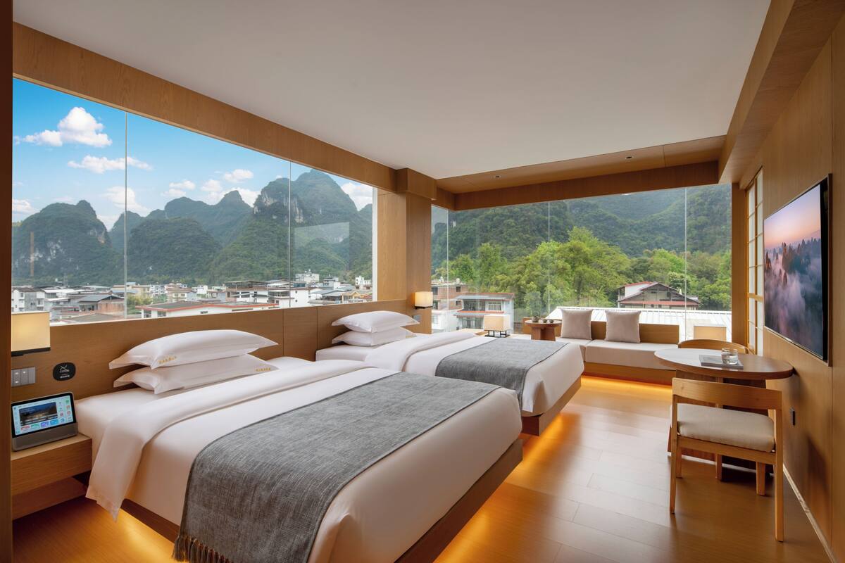Habitación de hotel con dos camas, un escritorio, una silla y un amplio ventanal con vistas a la montaña.