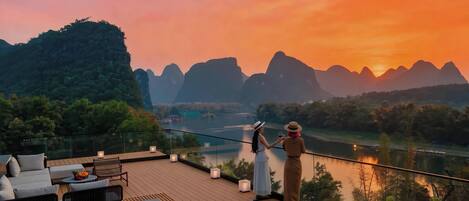 A terrace with a view of a river and mountains, furnished with sofas and tables.