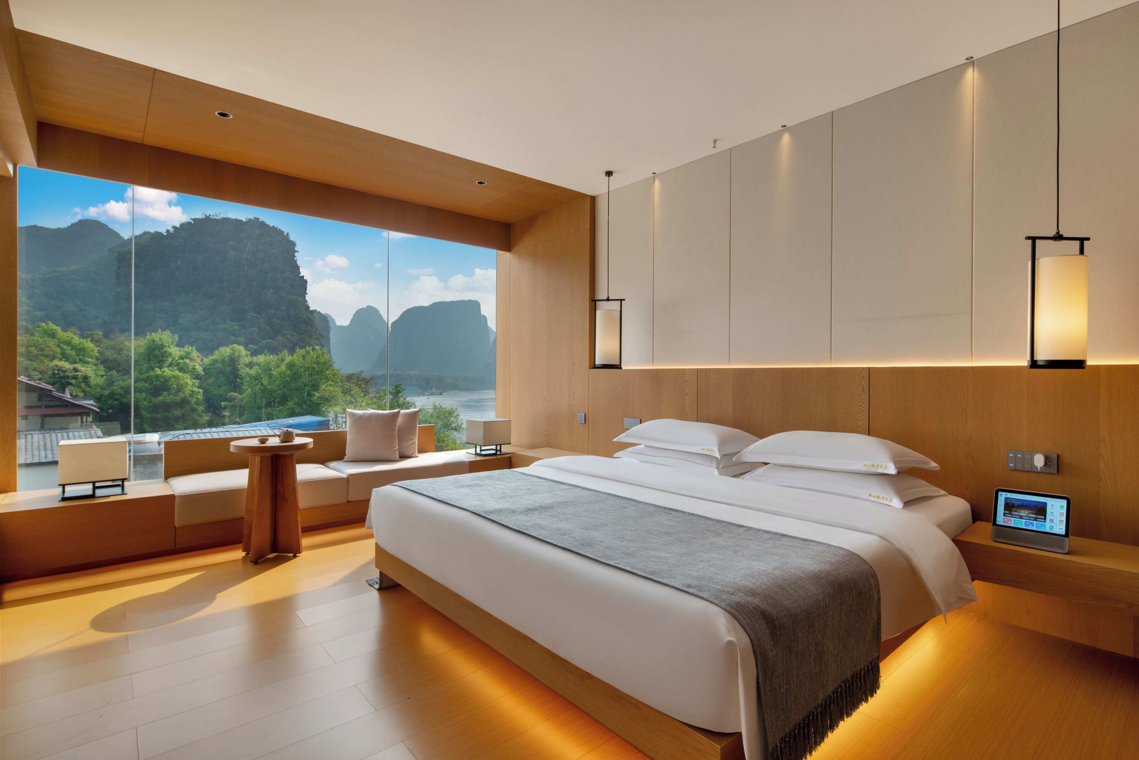 Un dormitorio moderno con una cama grande, una mesita de noche con un dispositivo digital, un sofá junto a la ventana y vistas a un paisaje montañoso.