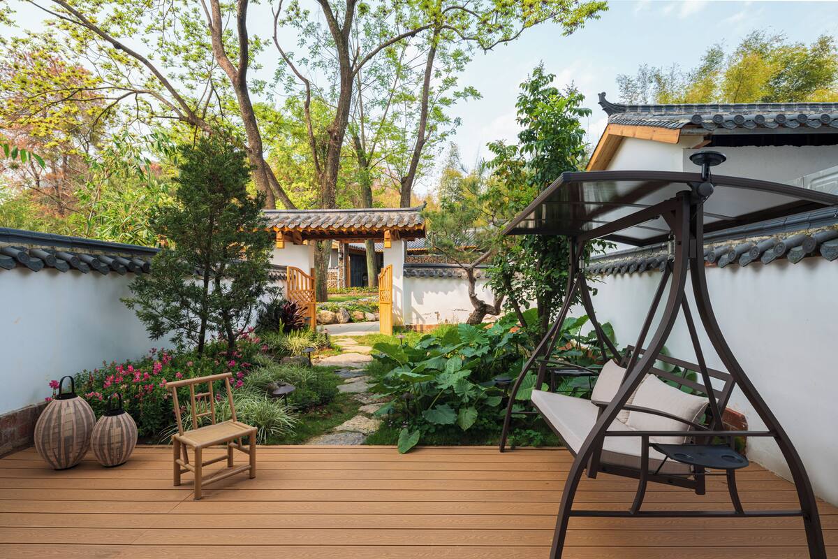 Una terraza de madera con una silla mecedora, un banco pequeño de madera y un edificio de estilo tradicional con tejado de baldosas.