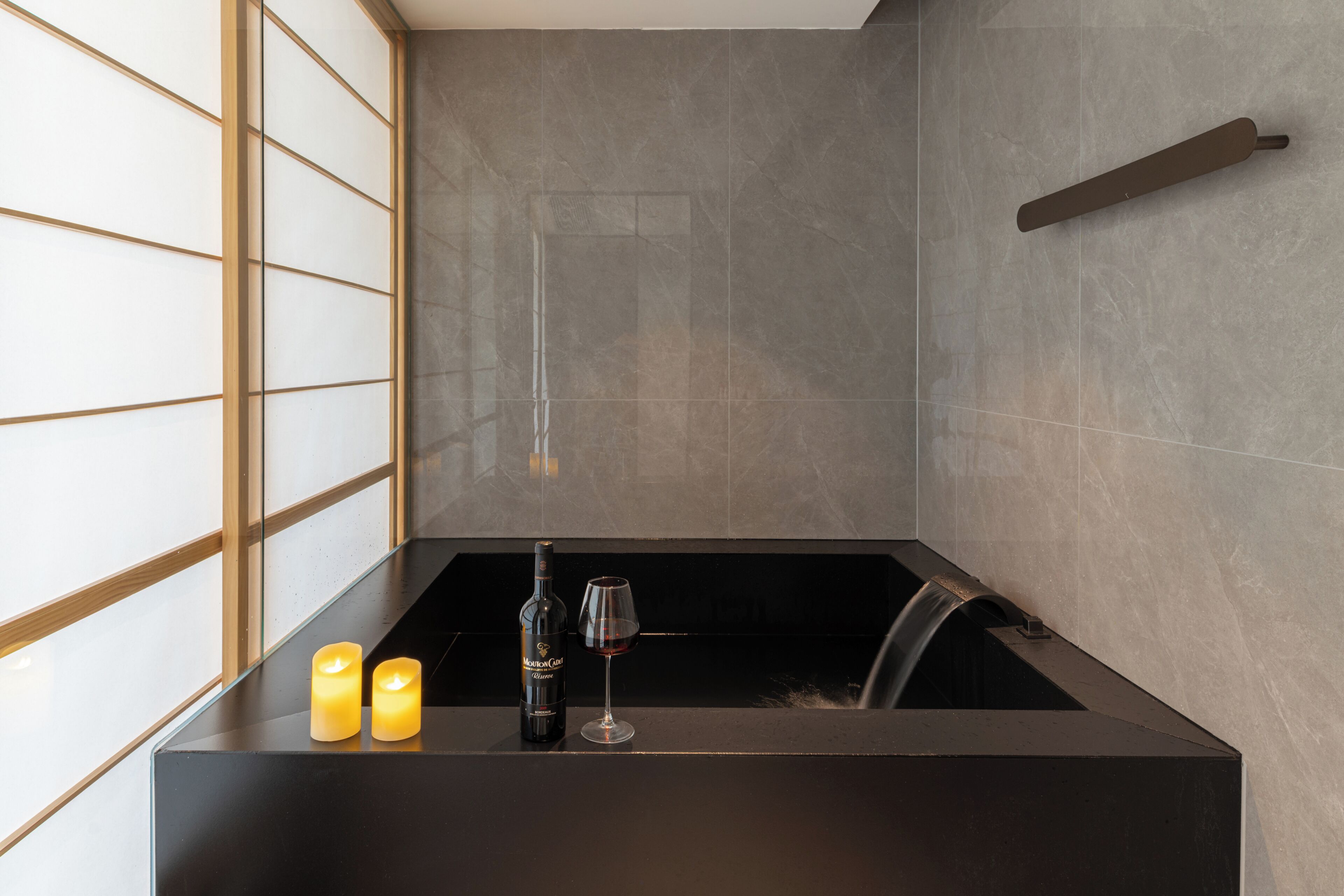 A modern bathroom with a black bathtub, a wine bottle, and two candles on a black countertop.