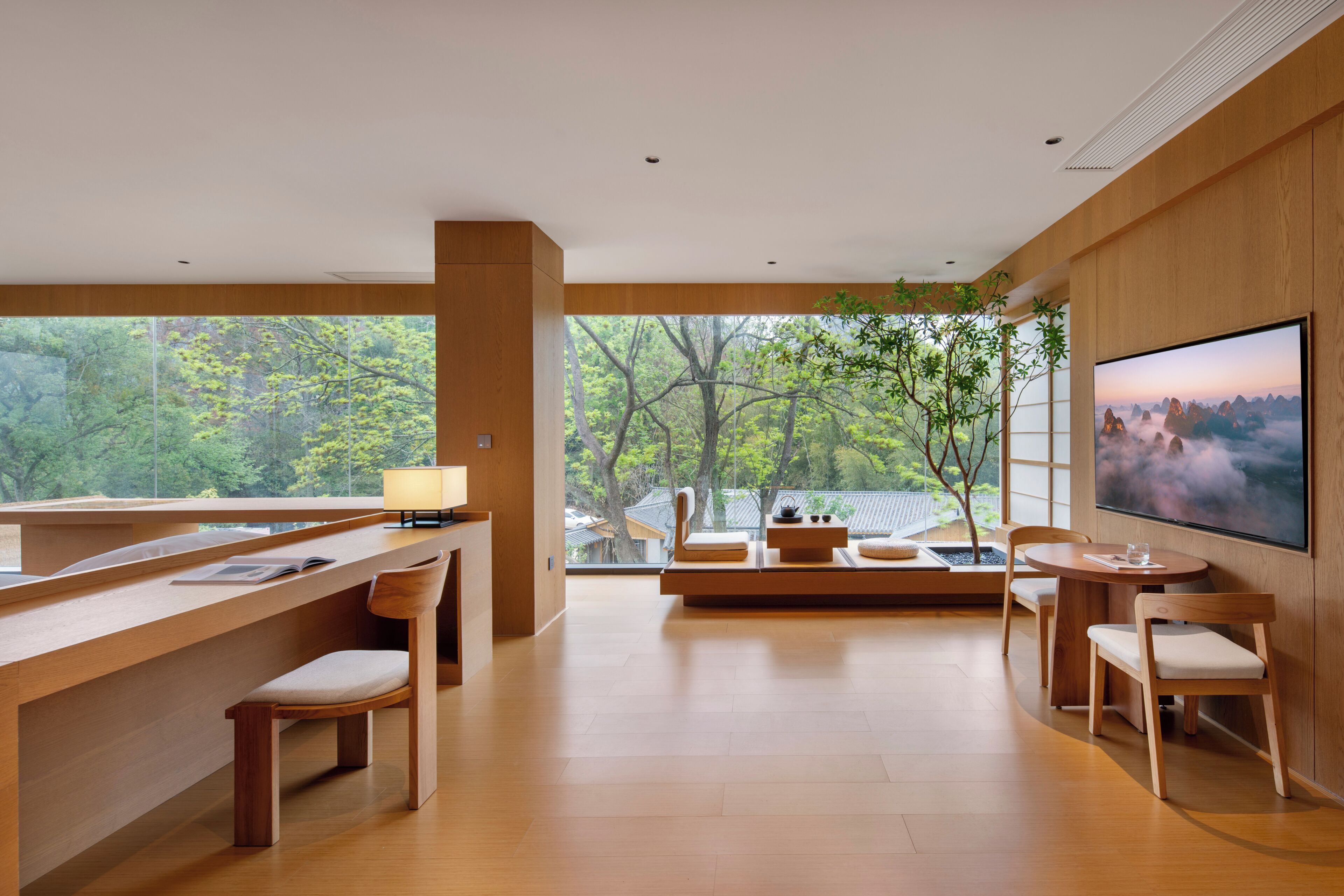 Un intérieur moderne avec un bureau en bois, des chaises, un téléviseur et une grande fenêtre donnant sur la forêt.