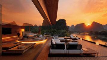 Un moderno espacio lounge al aire libre con terraza de madera, arreglos de asientos y vistas a un río al atardecer.