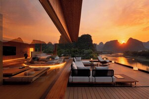 Un moderno espacio lounge al aire libre con terraza de madera, arreglos de asientos y vistas a un río al atardecer.