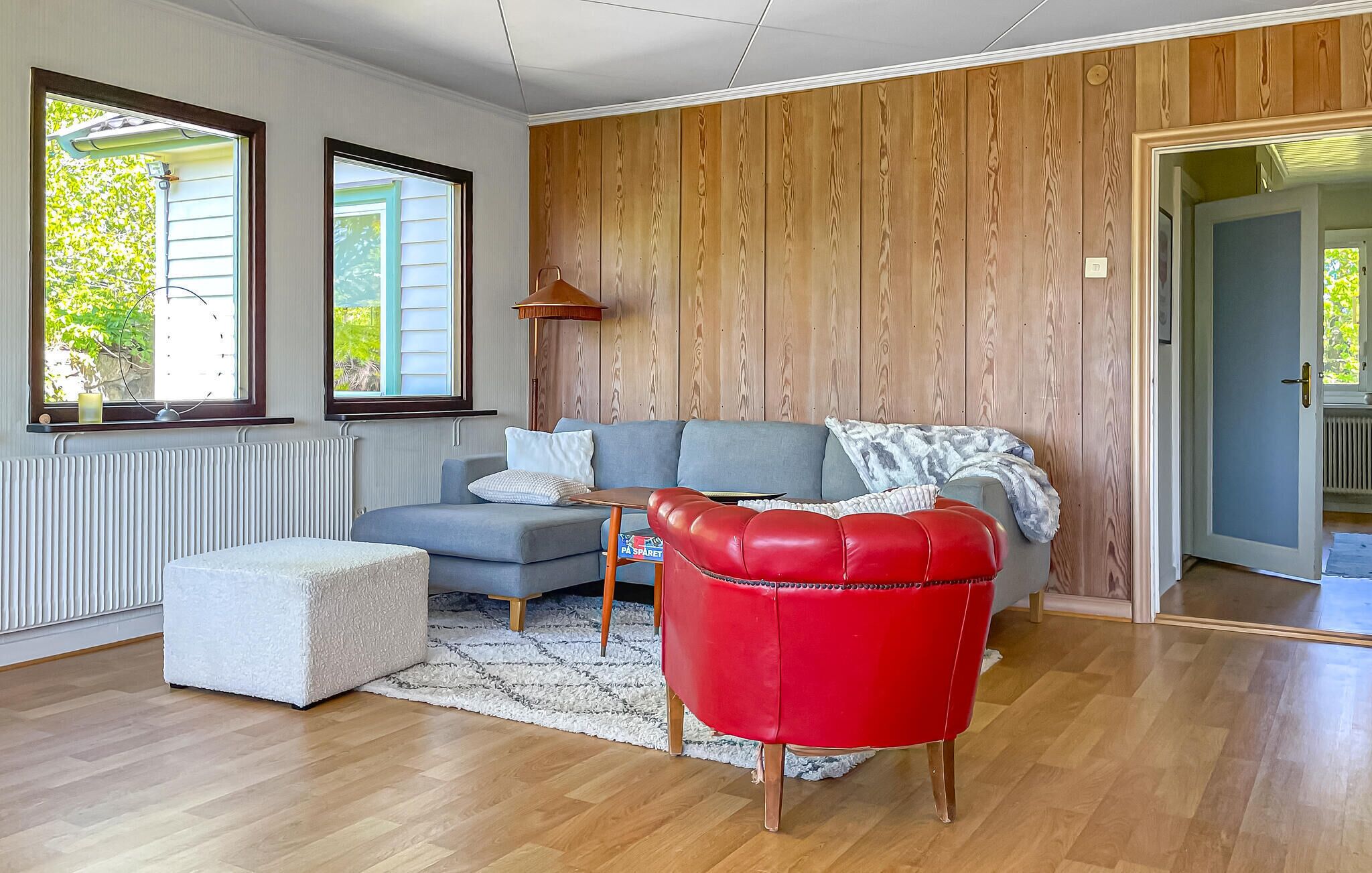 Uma sala de estar com um sofá cinzento, uma poltrona vermelha, um banquinho branco, painéis de parede de madeira e uma janela com persianas.