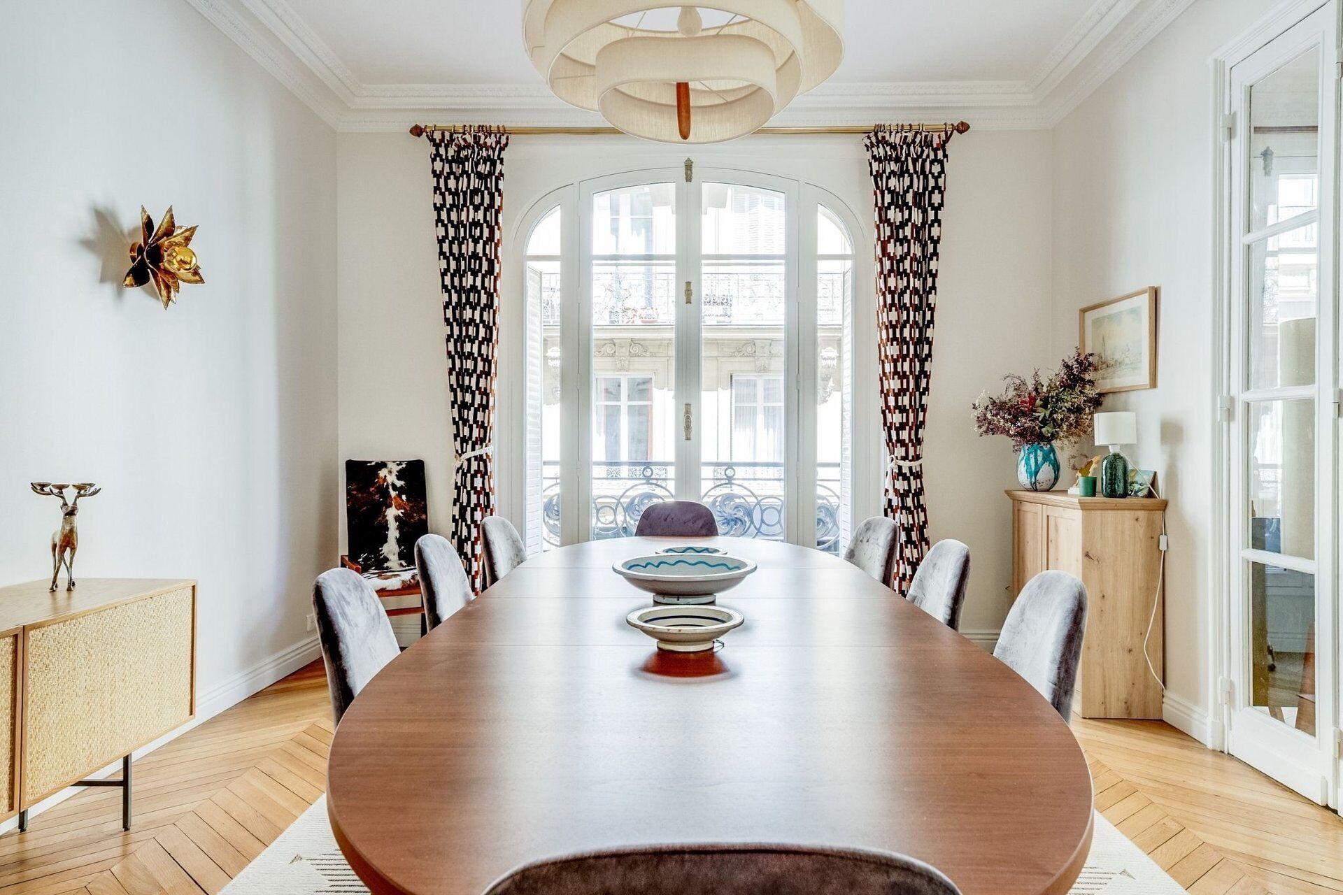 Un comedor con una gran mesa de madera, sillas, una araña y cortinas estampadas.