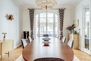A dining room with a large wooden table, chairs, a chandelier, and patterned curtains. - L'Ivoire – Elegant Family Haven-by Mon Petit Faré (Paris)
