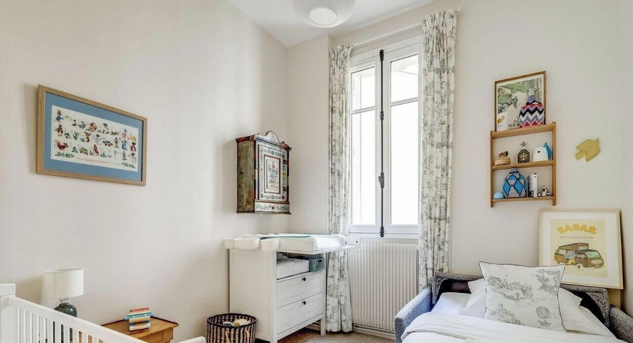 A bedroom with a crib, a dresser, a radiator, a window with curtains, and a bed with pillows.