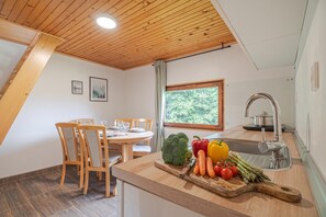 A modern kitchen with wooden cabinetry, a dining area with a round table and chairs, and a large window providing a view of greenery.
