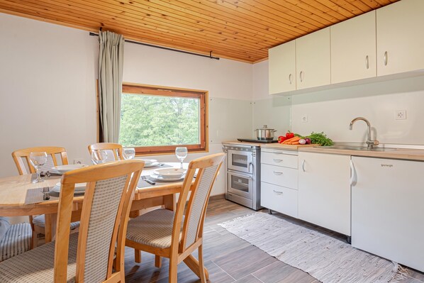 A modern kitchen with white cabinets, a built-in oven, and a dining area with wooden chairs and a table set for two. - Lodge Trtinka /w Terrace & BBQ in Nature (Zalec)