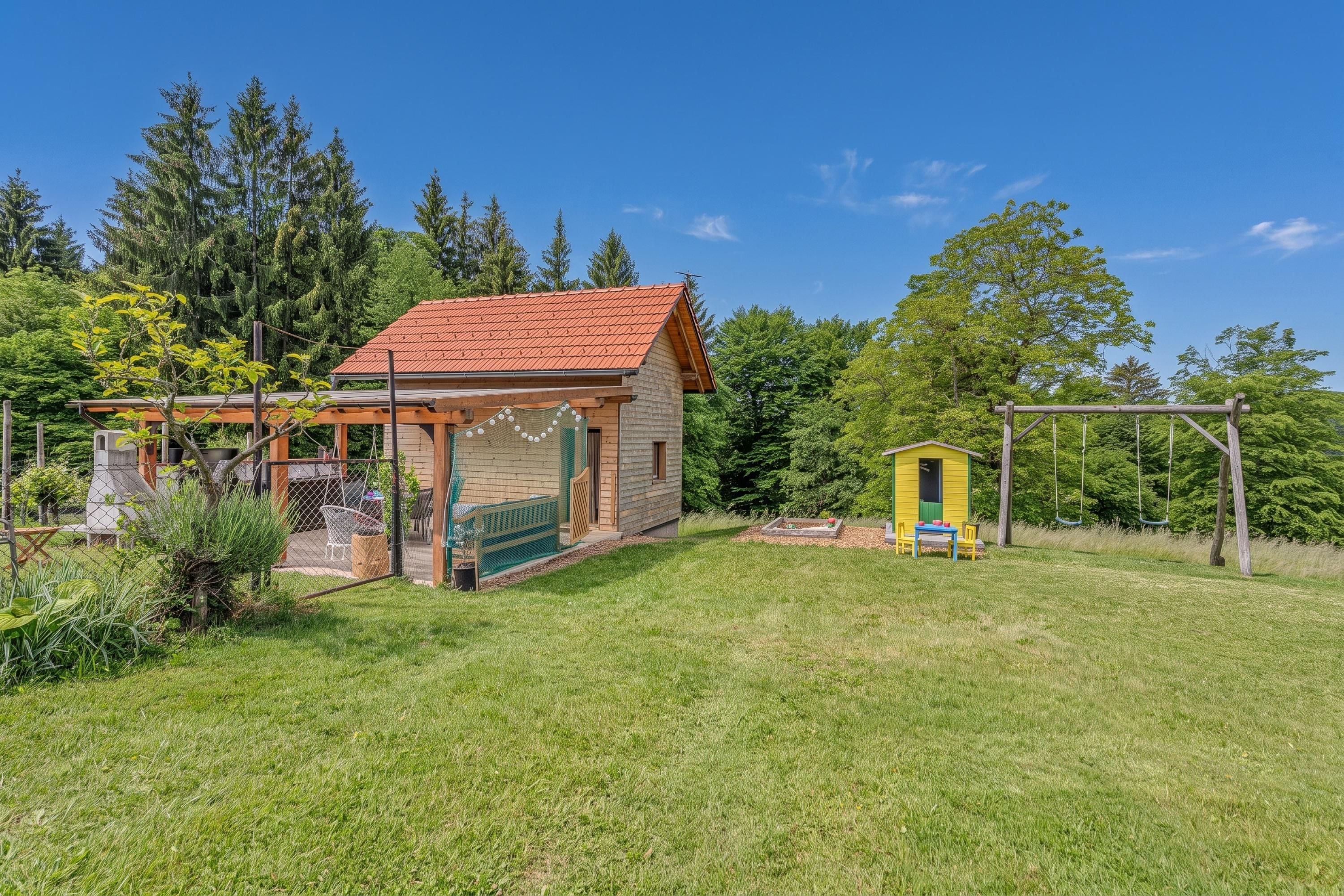 A small wooden house with a red roof, a fenced patio area, and a swing set.