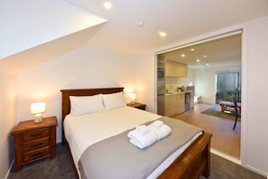 A bedroom with a bed, bedside tables, and a view of the kitchen and balcony.