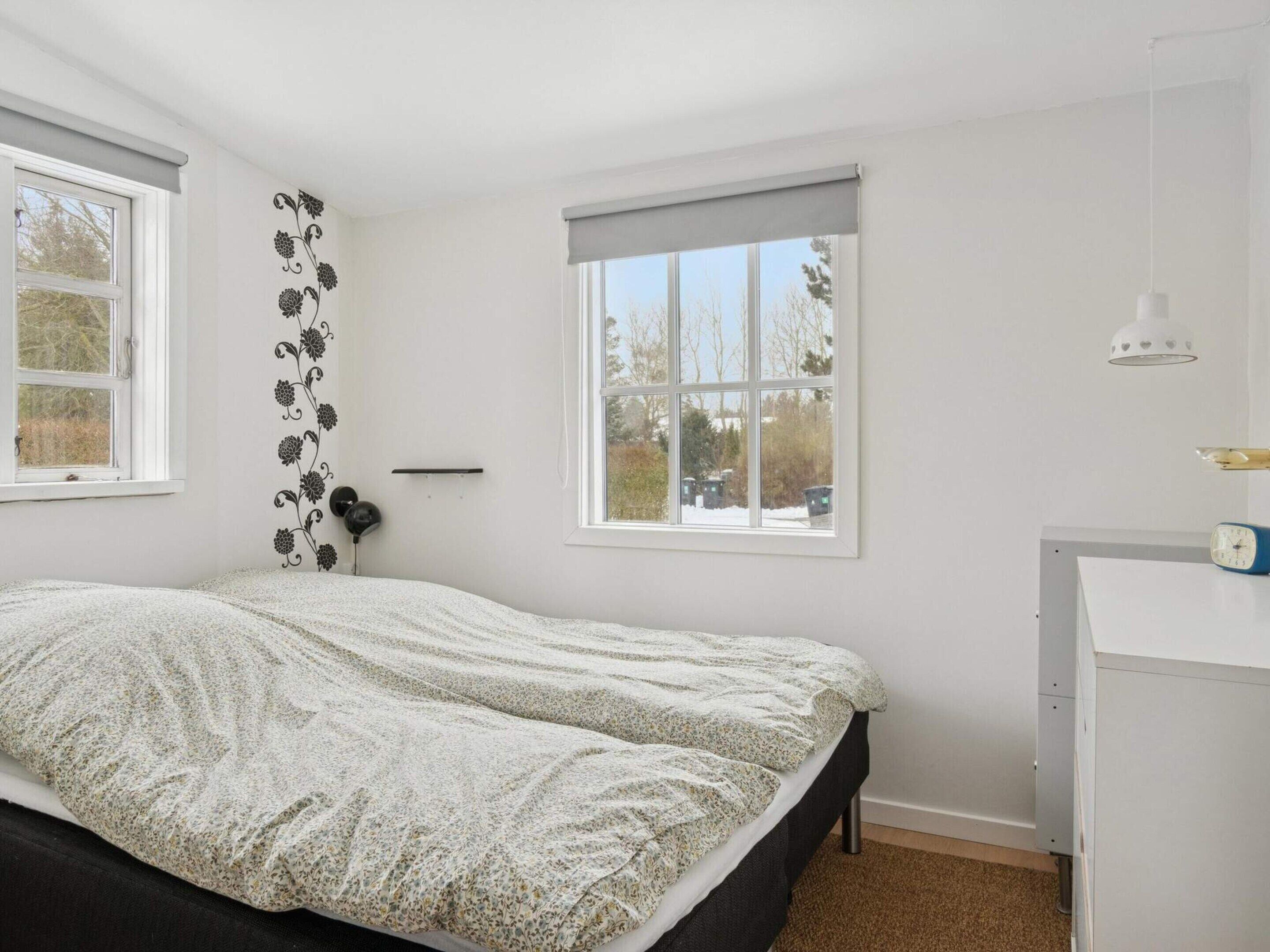 Um quarto com uma cama, duas janelas, uma mesa de cabeceira e uma escrivaninha com abajur.
