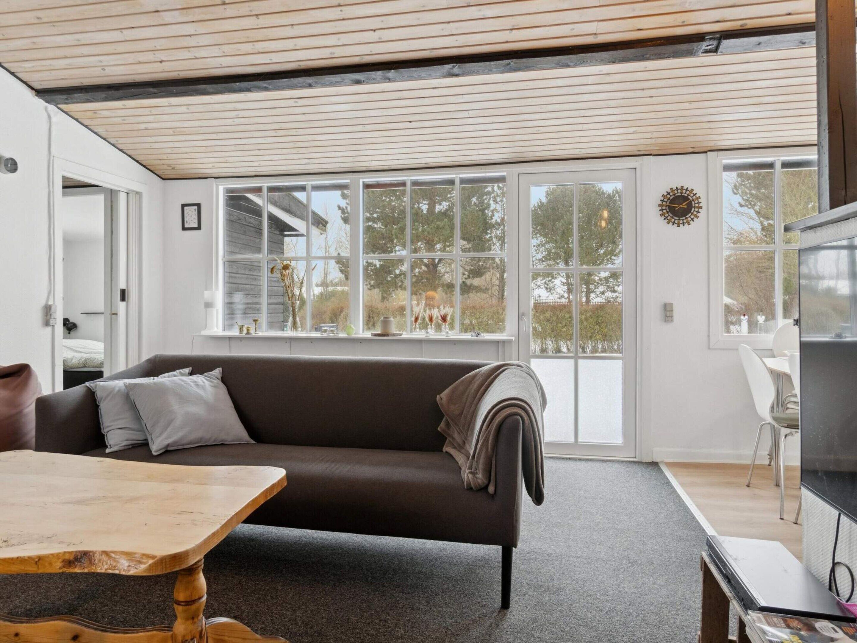 A modern living room with a sofa, a wooden table, and a TV. Large windows provide a view of the outdoors.