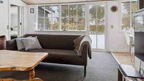 A modern living room with a sofa, a wooden table, and a TV. Large windows provide a view of the outdoors. - 4 Star Holiday Home in Nakskov (Nakskov)