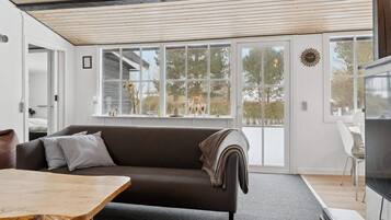 A modern living room with a sofa, a wooden table, and a TV. Large windows provide a view of the outdoors.