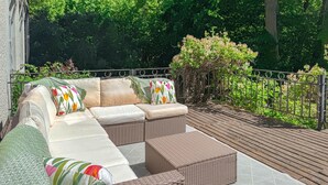 A covered outdoor seating area with a wicker sofa and cushions, surrounded by greenery and a wooden deck. - 4 bedroom stunning home in Borghamn (Borghamn)