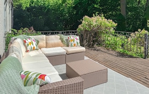 Ein überdachter Außenbereich mit Korbmöbel-Sofa und Kissen, umgeben von Grünpflanzen und einer Holzterrasse.