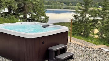 A hot tub with a wooden exterior, a built-in seat, and a step for access. It is situated on a gravel surface with a lake and forested mountains in the background.