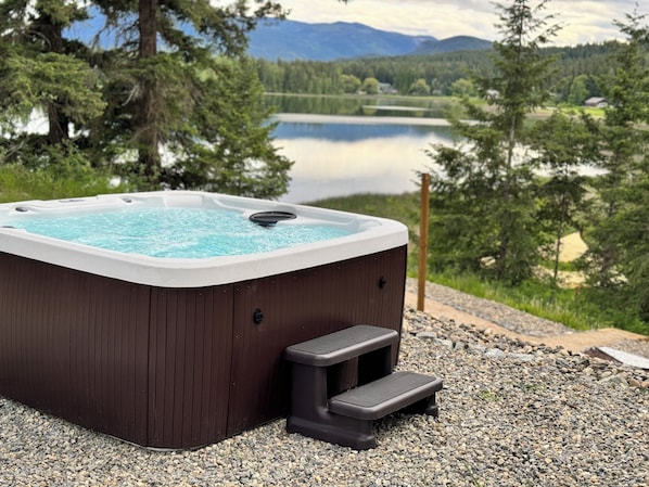Una vasca idromassaggio con rivestimento in legno, seduta integrata e un gradino per l'accesso. Si trova su una superficie sterrata, con un lago e montagne boscose sullo sfondo.
