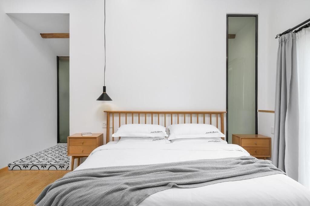 A modern bedroom with a wooden headboard, a single bed with white bedding, a bedside table, a nightstand, a hanging light fixture, and a large mirror.