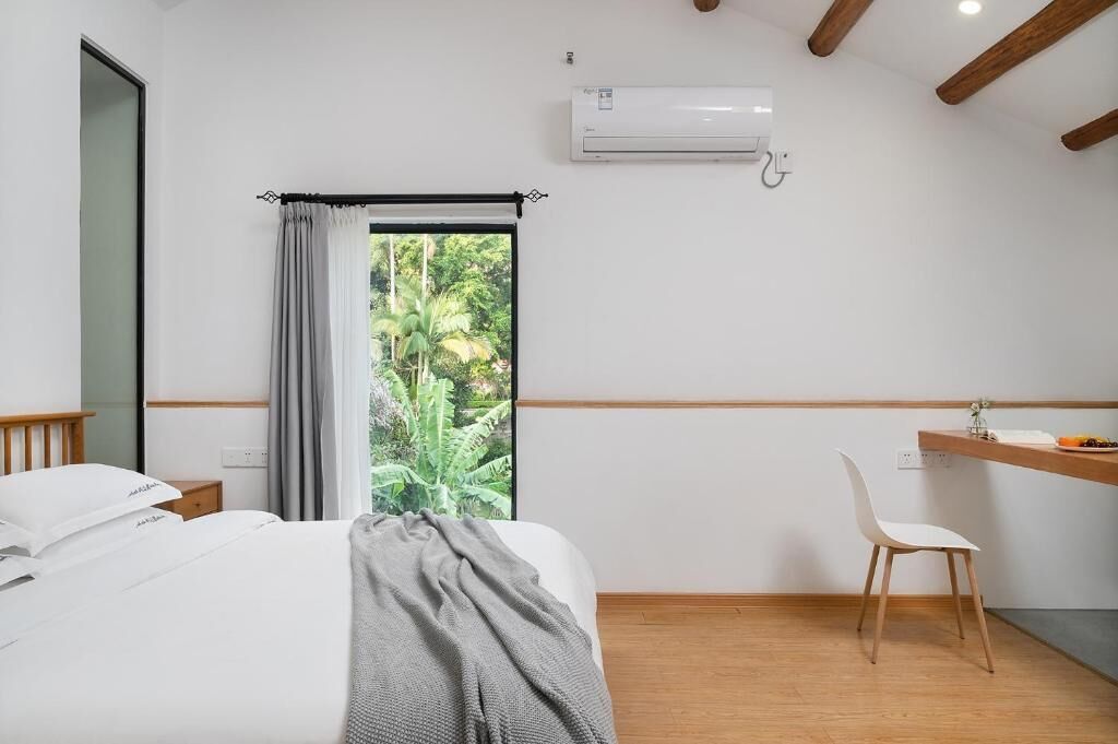A bedroom with a bed, a desk, a chair, a window with curtains, an air conditioner, and a view of greenery.