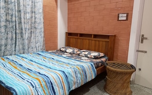 A single bed with a patterned bedspread, a wooden headboard, a wall-mounted light switch, and a door with a handle.