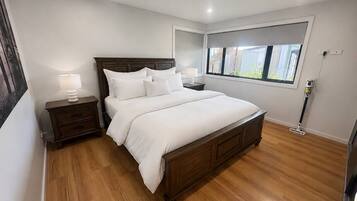 A bedroom with a wooden bed, a nightstand with a lamp, a window with a view of buildings, and a ceiling light.