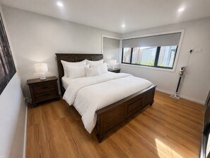 Ein Schlafzimmer mit einem hölzernen Bett, einem Nachttisch mit Lampe, einem Fenster mit Blick auf Gebäude und einer Deckenleuchte.