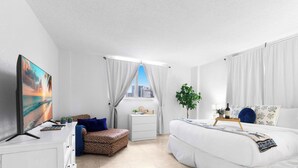 A modern hotel room with a bed, a desk, a chair, a window with curtains, a plant, and a city view. - Oceanview Comfort Elegant S Ocean Drive (Hollywood)