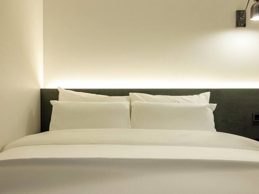 A neatly made bed with white linens, a dark headboard, and a wall-mounted light fixture.