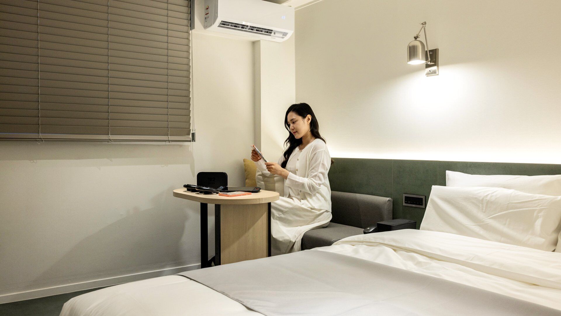 Habitación de hotel con cama, mesita de noche con lámpara, ventana con persianas y equipo de aire acondicionado.