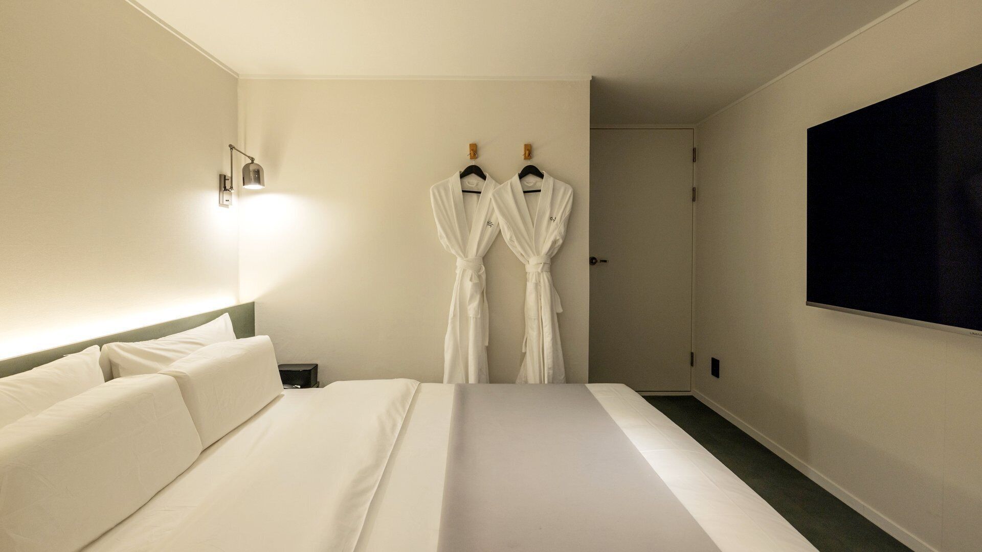 Habitación de hotel con una cama, dos batas colgadas en ganchos, una lámpara de pared y un televisor en la pared.