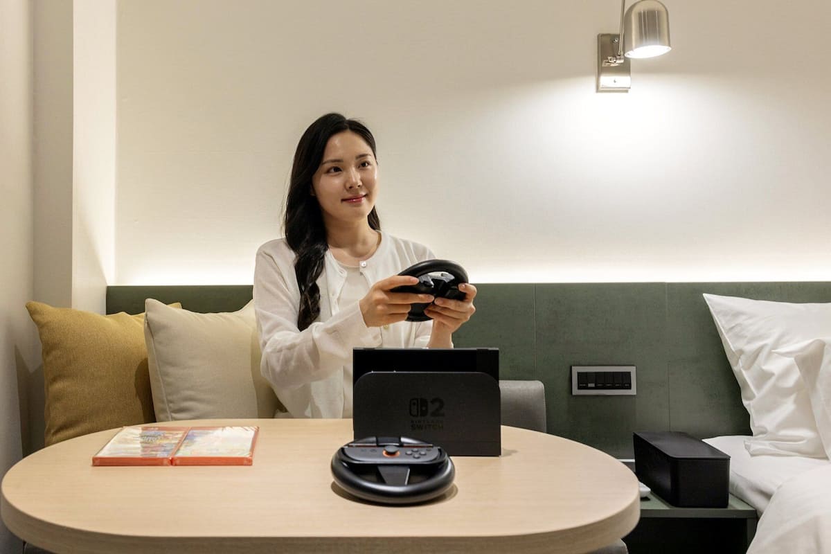 A person seated at a round table in a hotel room, holding a game controller.