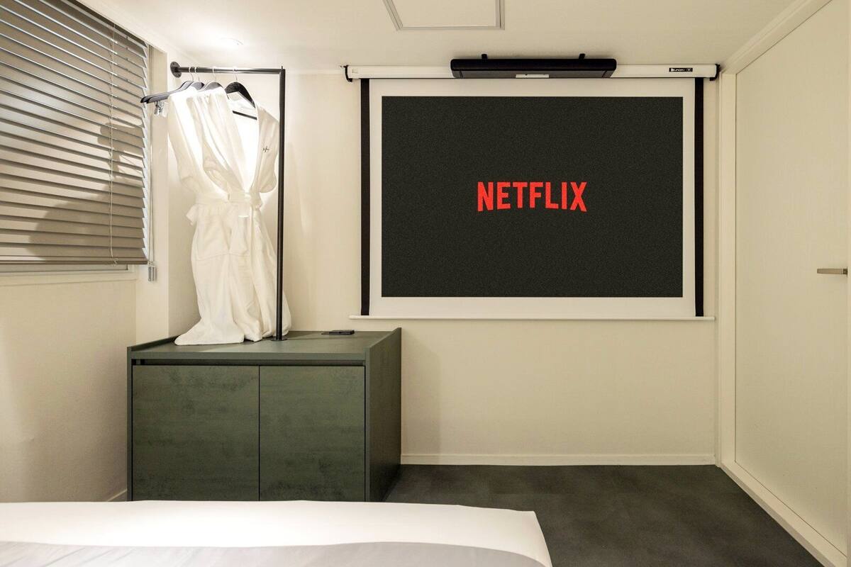 A bedroom with a bed, a small dresser, a wall-mounted projector screen displaying 'NETFLIX,' and a window with blinds.