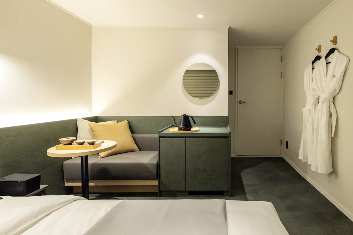 A hotel room with a bed, a sofa, a small table with a kettle, a robe hanging on the wall, and a door.
