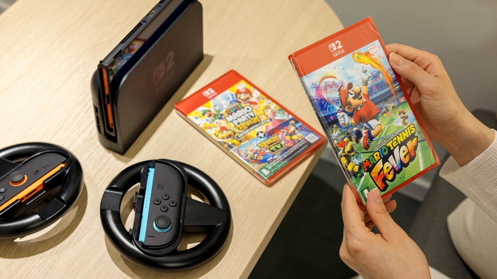 Eine Person hält einen Mario Kart Spielekoffer, eine Nintendo Switch Konsole und zwei Lenkräder auf einem Holztisch.