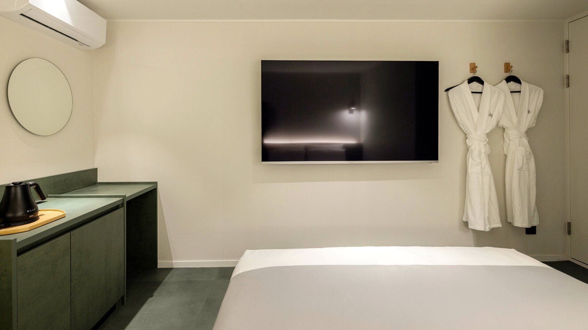 A hotel room with a bed, a flat-screen TV, a coffee maker, a round mirror, and two white robes hanging on a door.