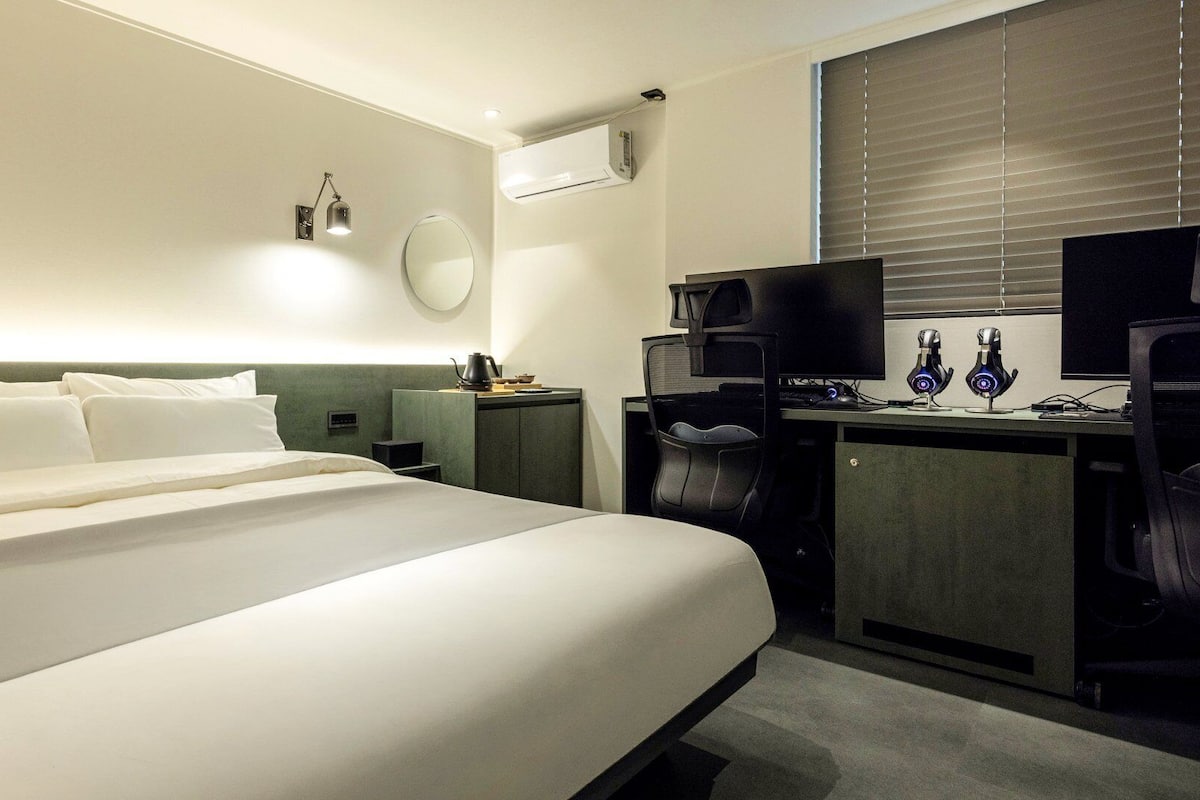A hotel room with a large bed, a desk with a computer and two webcams, a round mirror, and an air conditioning unit.
