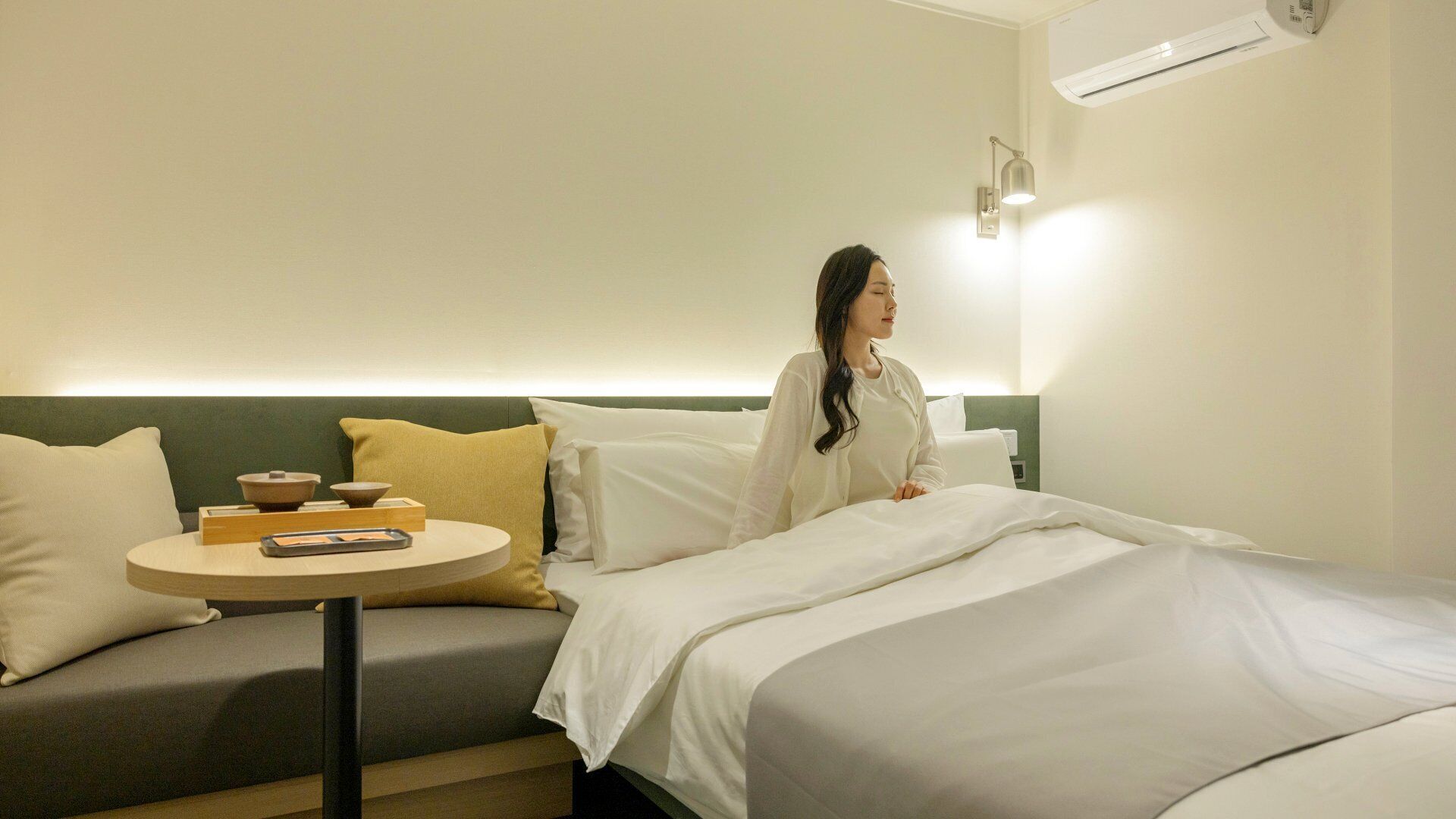 A hotel room with a bed, a small round table with a remote control, and a wall-mounted air conditioner.