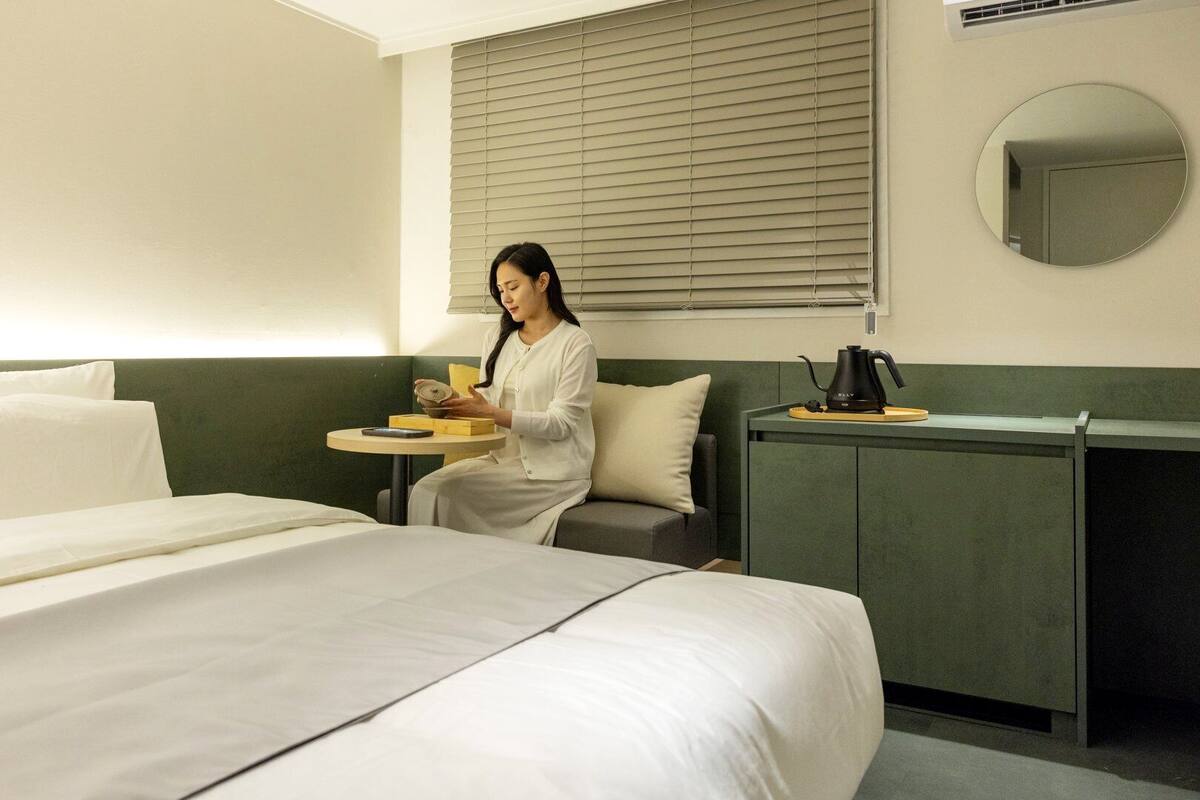 A hotel room with a bed, a bedside table, a chair, a small table with a teapot, and a window with blinds.