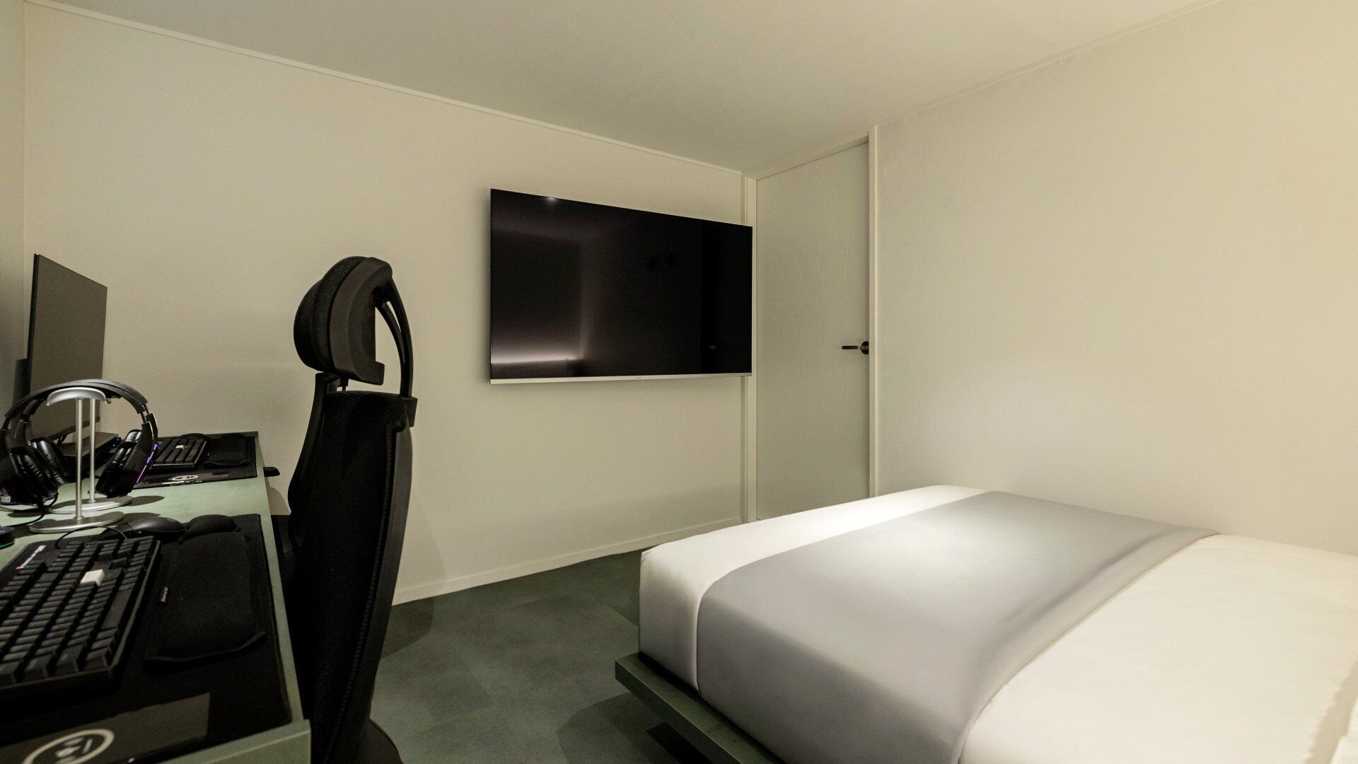A hotel room with a bed, a desk with a computer, headphones, and a TV mounted on the wall.