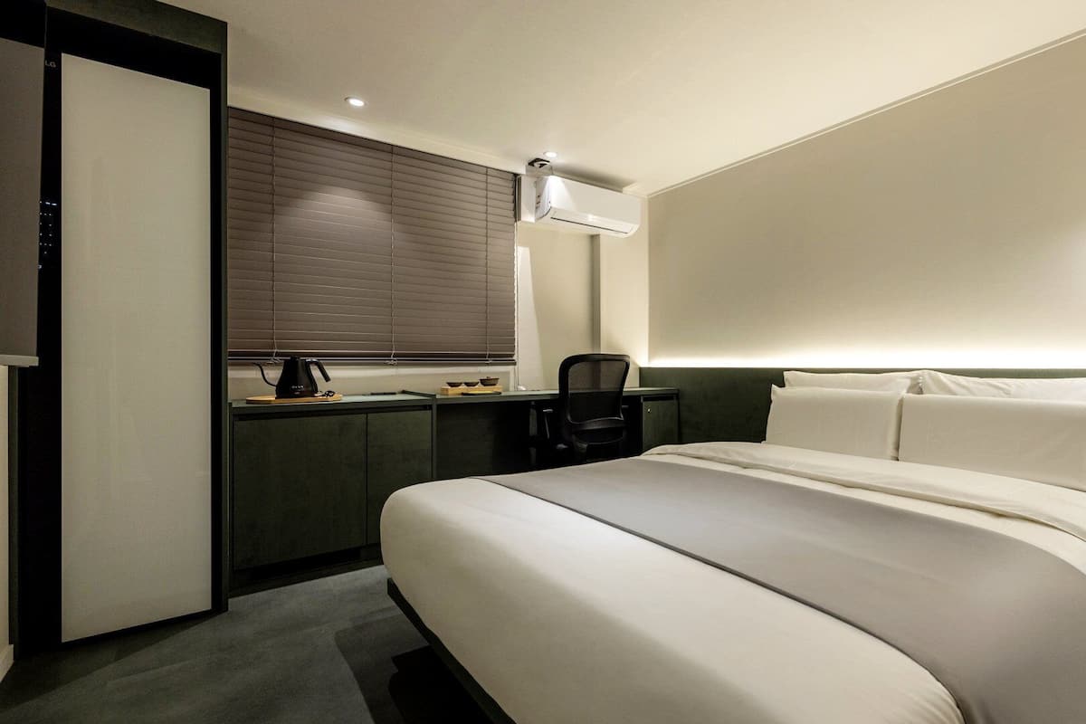 A hotel room with a bed, a desk, a chair, a coffee maker, and a wall-mounted light.